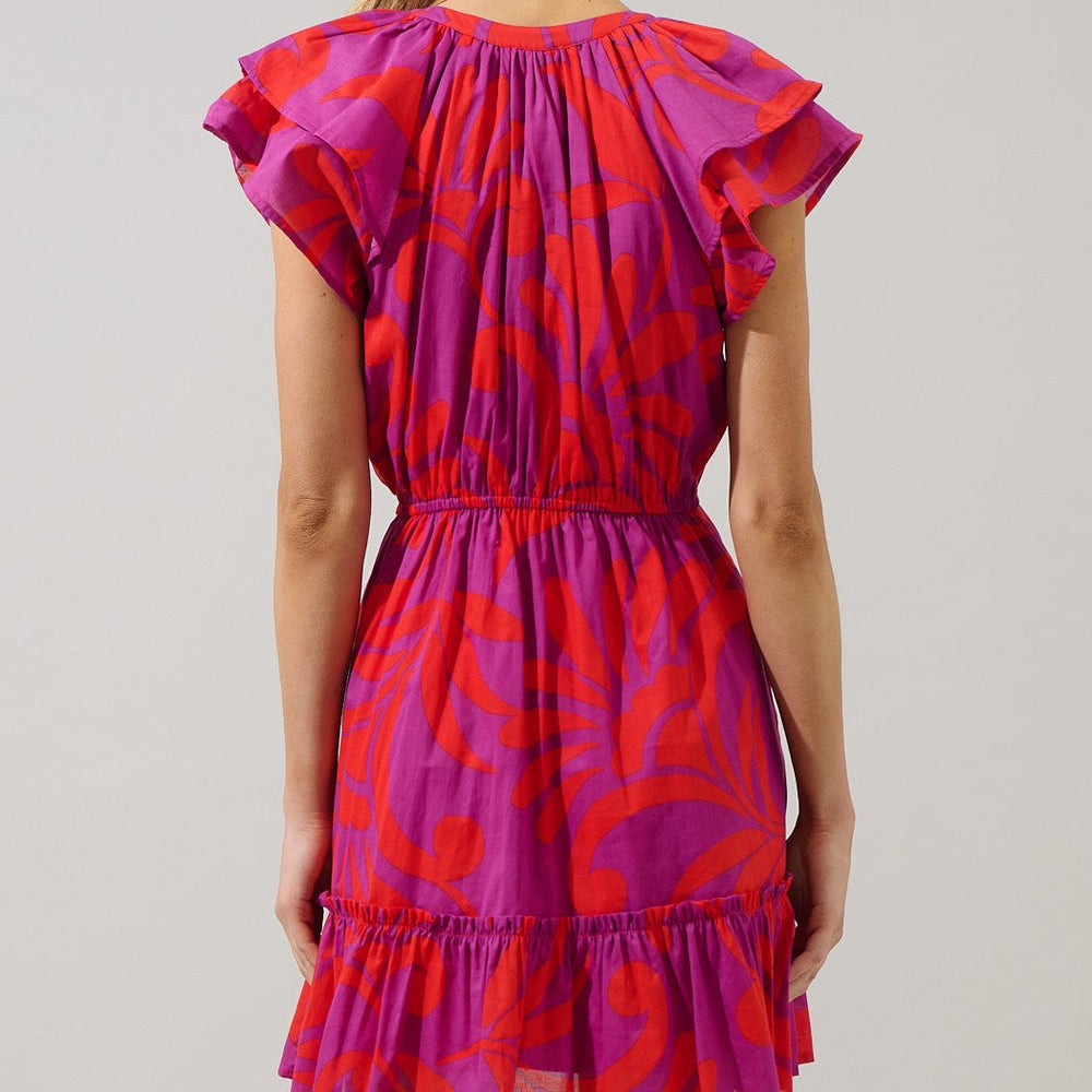 Digital photograph of a woman from behind in a white studio wearing a short dress with a bold magenta and orange abstract pattern featuring layered flutter sleeves and a cinched elastic waist