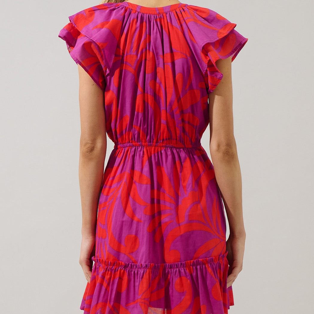 Digital photograph of a woman from behind in a white studio wearing a short dress with a bold magenta and orange abstract pattern featuring layered flutter sleeves and a cinched elastic waist