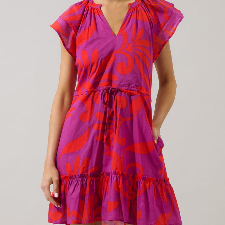 Studio fashion photograph of a woman modeling a bold purple and red floral print dress in a minimalist studio setting featuring a cinched drawstring waist, flutter sleeves, and a ruffled tiered hem