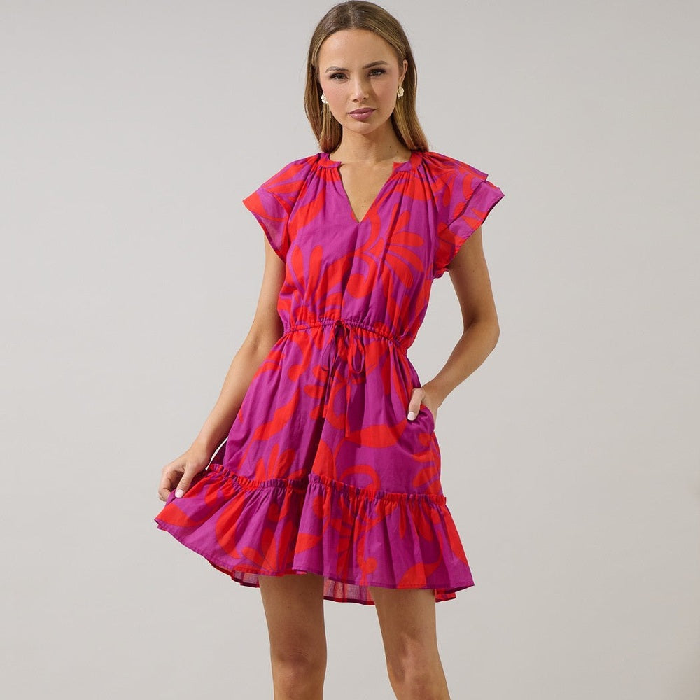 Studio photograph of a blonde model wearing a vibrant magenta and orange abstract leaf print mini dress with flutter sleeves, a drawstring waist, and a ruffled hem against a neutral grey background
