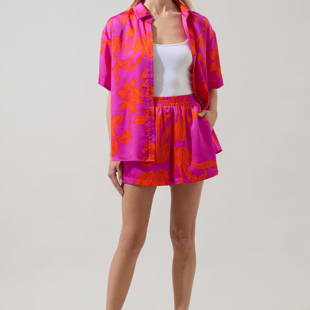 Studio photograph of a model in a neutral studio wearing a vibrant hot pink and orange tropical floral print satin shirt and matching shorts set layered over a white tank top