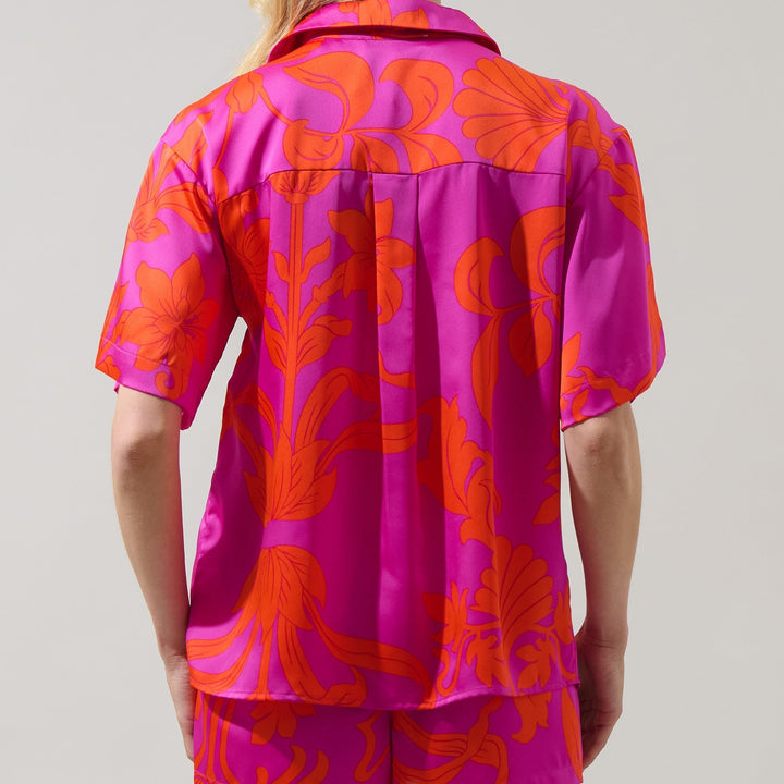 Studio photograph of a person from behind in a neutral setting wearing a vibrant fuchsia pink short-sleeved shirt and matching shorts featuring a large-scale orange botanical floral print