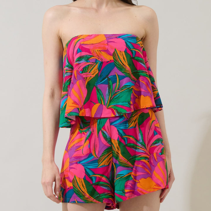 Fashion studio photograph of a model wearing a vibrant tropical two-piece set featuring a tiered strapless crop top and high-waisted shorts with a bold pattern of hot pink, orange, and teal foliage against a neutral wall