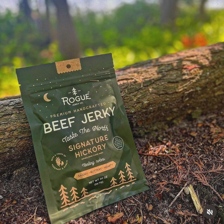 Digital photograph of Rogue Jerky Co. Signature Hickory beef jerky leaning against a fallen log in a sun-dappled forest featuring a dark green bag with a gold crescent moon and cream pine tree graphics