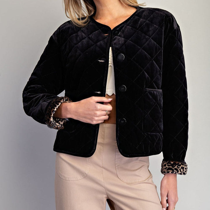 Studio photograph of a woman wearing a black diamond-quilted velvet jacket with leopard print sleeve lining and large buttons paired with beige trousers against a neutral backdrop