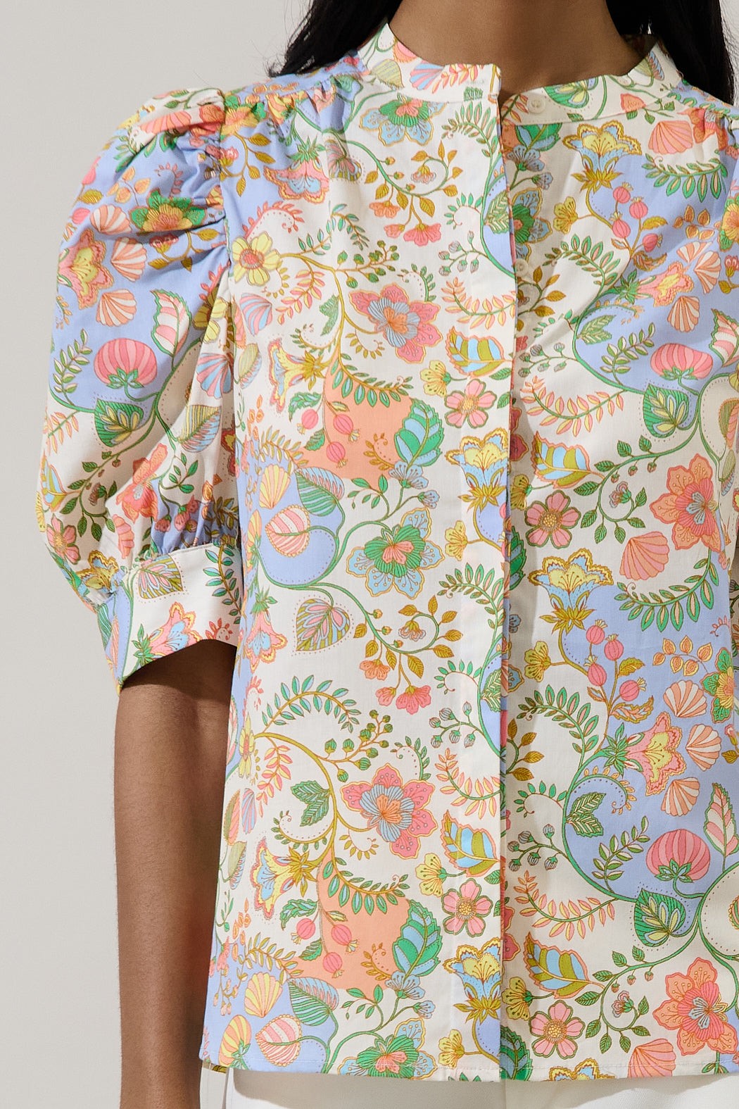 Cotton poplin blouse featuring a whimsical pastel botanical print with peach and mint green accents worn by a model in a studio setting highlighting voluminous cuffed puff sleeves and a button-front high collar