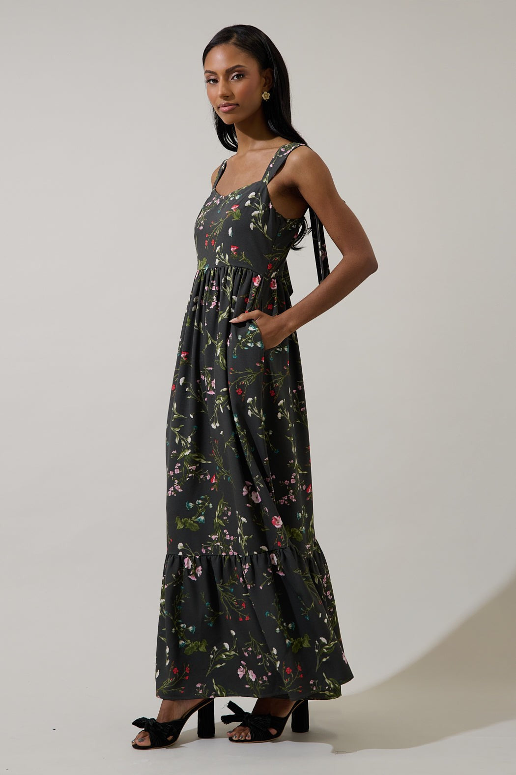 A studio photograph of a model in a dark floral-print maxi dress with a tiered hem, shoulder ties, and side pockets standing against a neutral background wearing black knotted heels and gold floral earrings