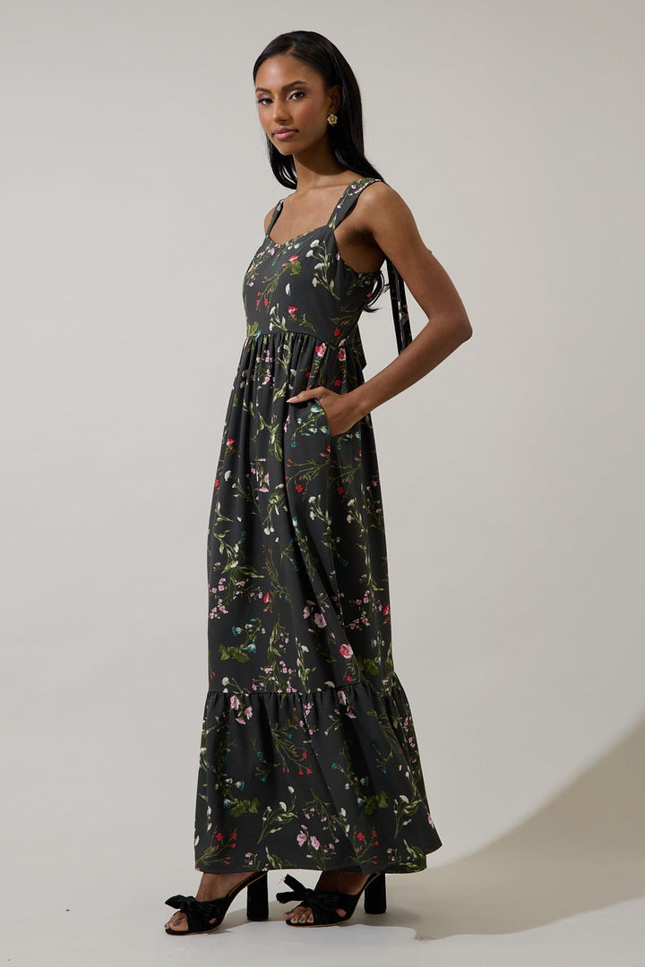 A studio photograph of a model in a dark floral-print maxi dress with a tiered hem, shoulder ties, and side pockets standing against a neutral background wearing black knotted heels and gold floral earrings