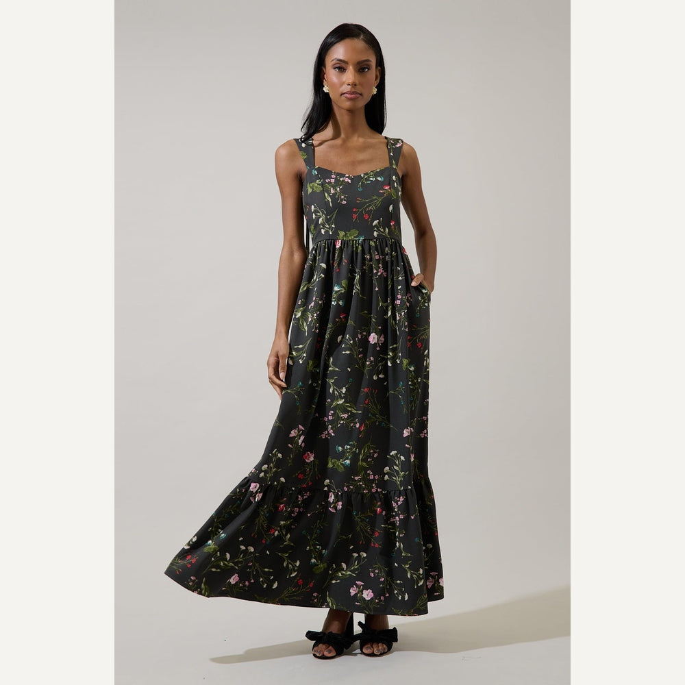 Fashion photograph of a woman standing in a neutral studio wearing a black floral maxi dress featuring a vibrant pink wildflower print, a tiered ruffled hem, and black bow sandals