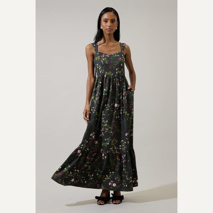 Fashion photograph of a woman standing in a neutral studio wearing a black floral maxi dress featuring a vibrant pink wildflower print, a tiered ruffled hem, and black bow sandals