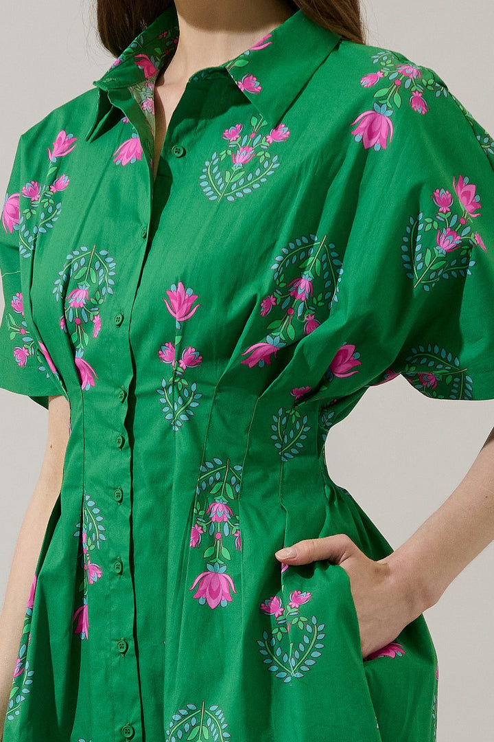 A fashion photograph of a woman in a studio wearing a vibrant emerald green button-down dress featuring pink floral lotus motifs, a cinched pleated waist, and light blue leafy accents