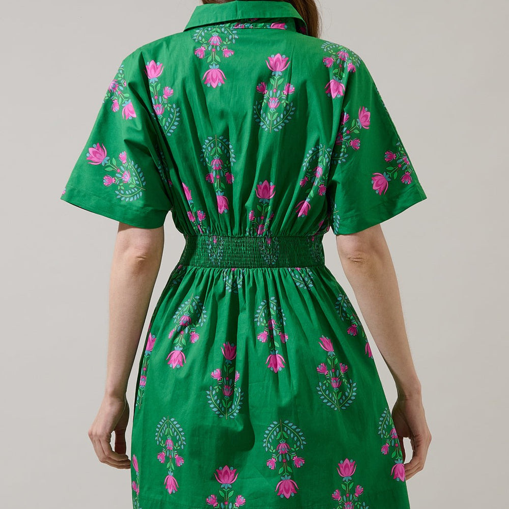 Digital photograph of a woman's back in a studio wearing a vibrant green short-sleeved dress featuring a pink floral lotus pattern and a smocked elastic waistline