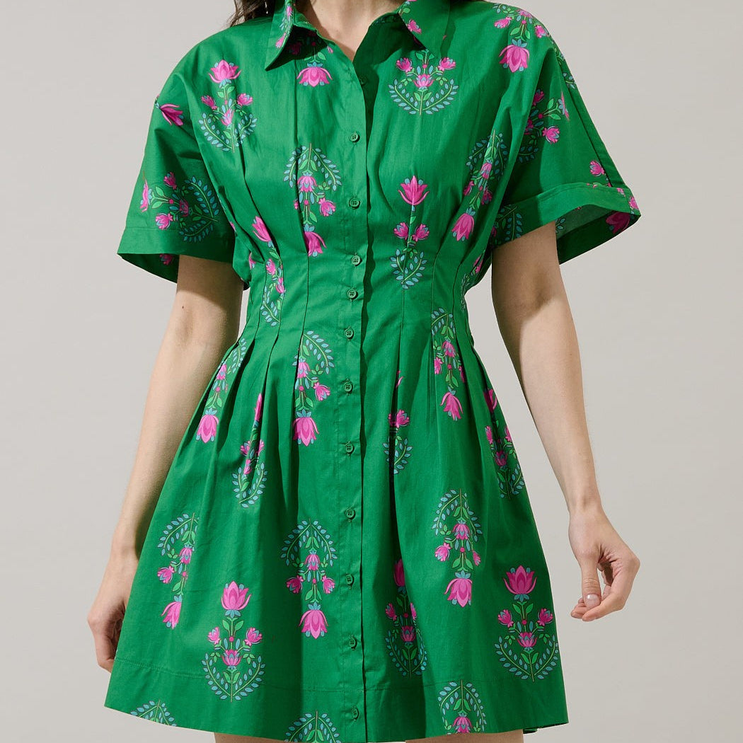 Studio photograph of a model wearing a vibrant green button-down shirt dress featuring bright pink floral motifs and a cinched pleated waistline against a neutral background