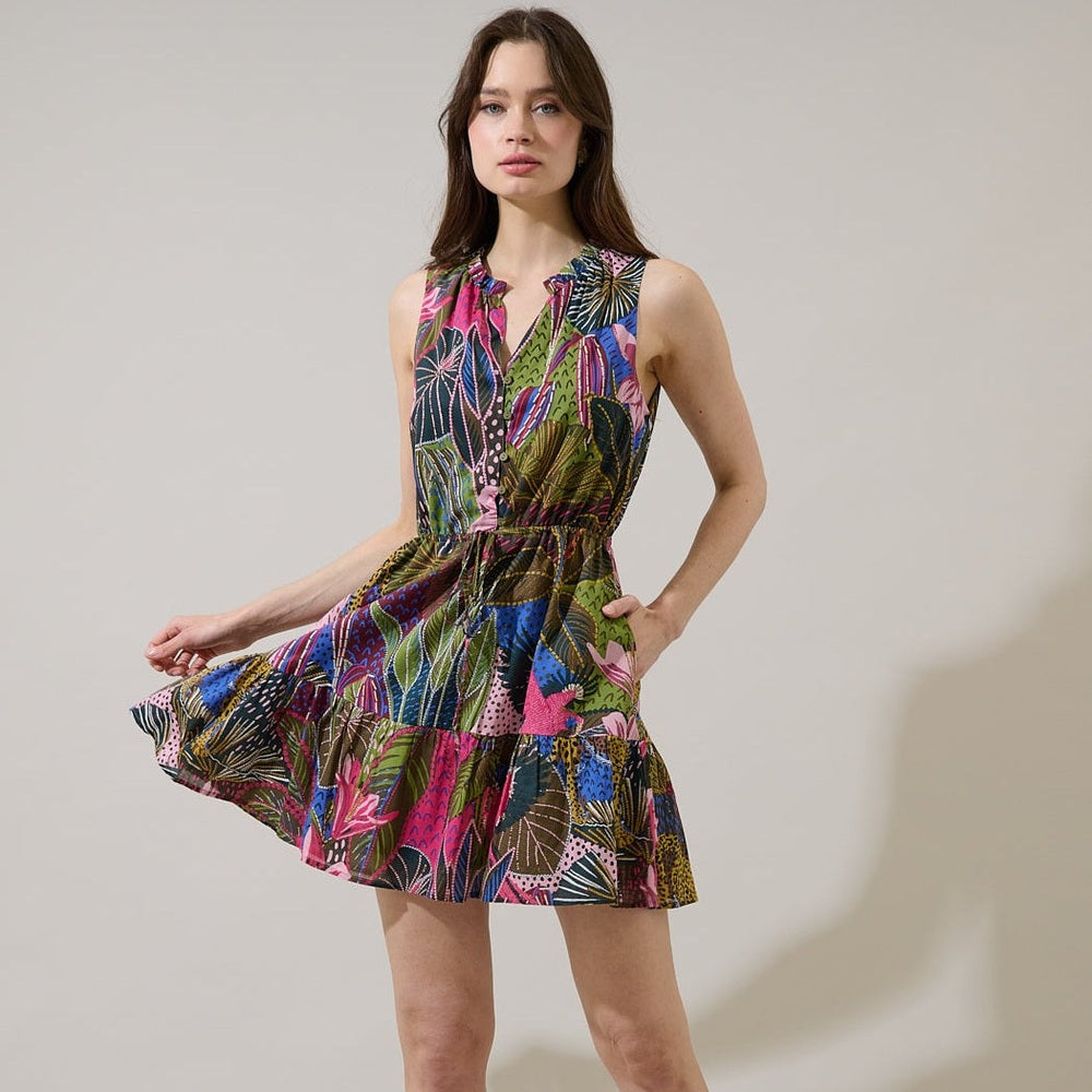 Fashion photograph of a woman in a minimalist studio wearing a vibrant botanical print mini dress featuring hot pink tropical leaves, a tiered ruffle skirt, and a cinched drawstring waist