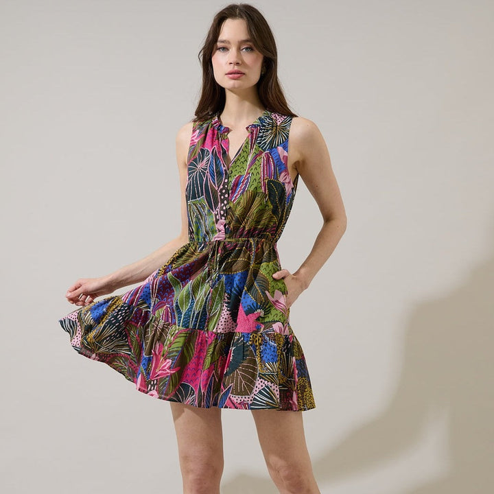 Fashion photograph of a woman in a minimalist studio wearing a vibrant botanical print mini dress featuring hot pink tropical leaves, a tiered ruffle skirt, and a cinched drawstring waist