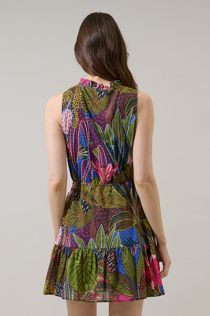 Fashion photograph of a woman from behind wearing a vibrant tiered dress with a tropical botanical print in a studio setting showcasing lush green leaves, pink floral accents, and stylized leopard patterns