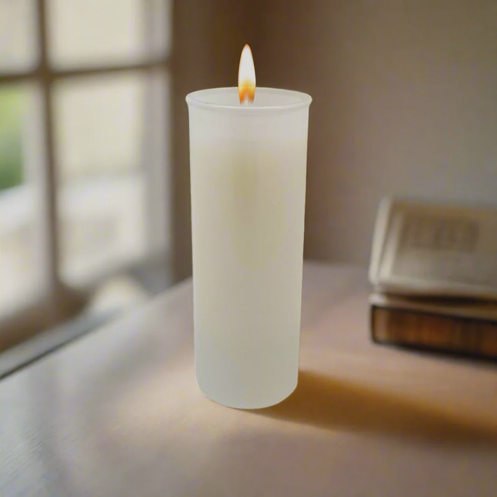 Digital photograph of a lit white pillar candle in a frosted glass holder on a table featuring a warm glowing flame, a blurred window frame, and a stack of antique books