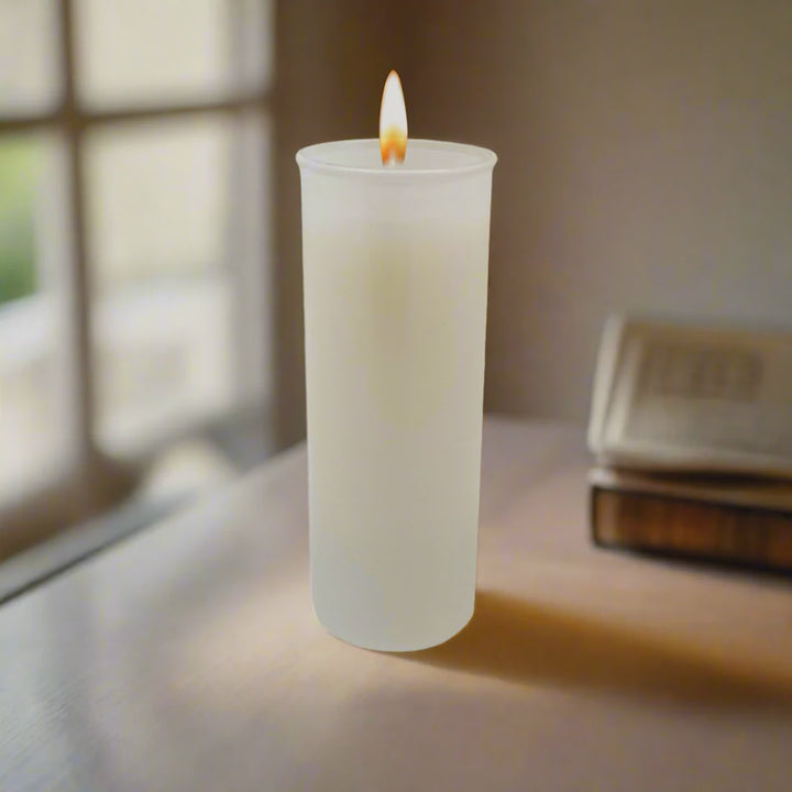 Digital photograph of a lit white pillar candle in a frosted glass holder on a table featuring a warm glowing flame, a blurred window frame, and a stack of antique books