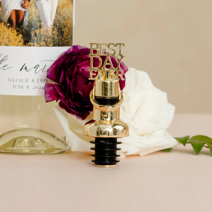 Photograph of a gold wine stopper featuring glittery Best Day Ever lettering positioned beside a personalized wedding wine bottle, a vibrant magenta ranunculus, and a soft white rose