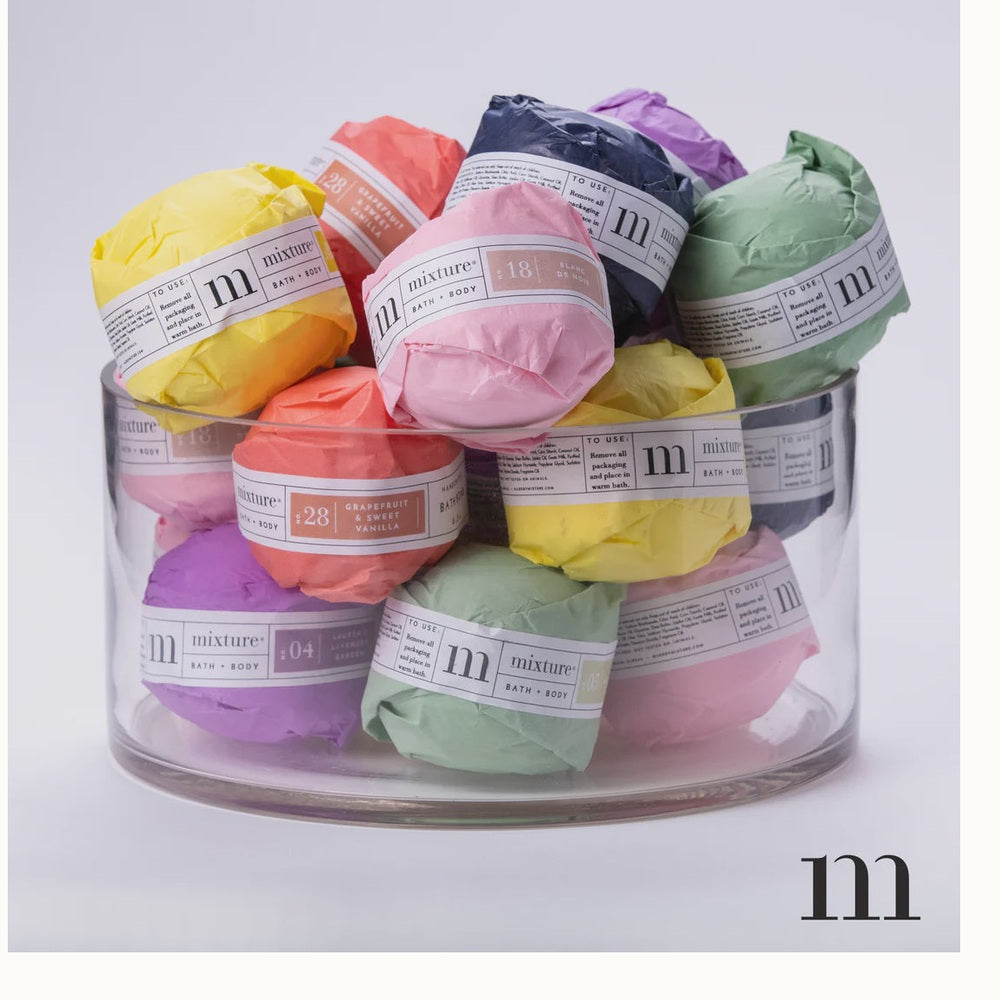 Studio photography by Mixture featuring a clear glass cylinder filled with bath bombs wrapped in colorful yellow, pink, and mint green tissue paper with white branding and the iconic black m logo against a white background