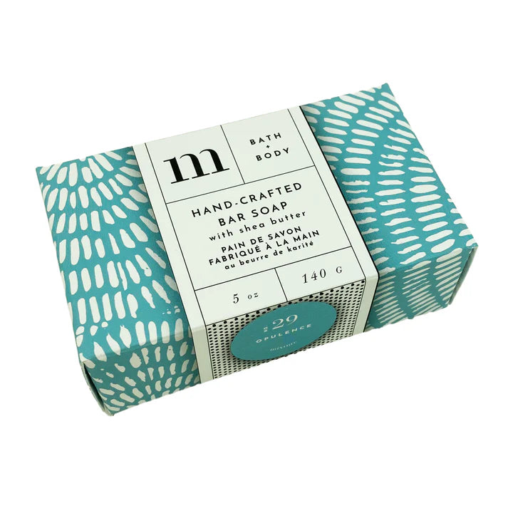 Product photography by Michaud of a hand-crafted bar soap in a teal and white dashed box centered on a white background featuring a minimalist paper wrap with a black m logo and a turquoise Opulence sticker