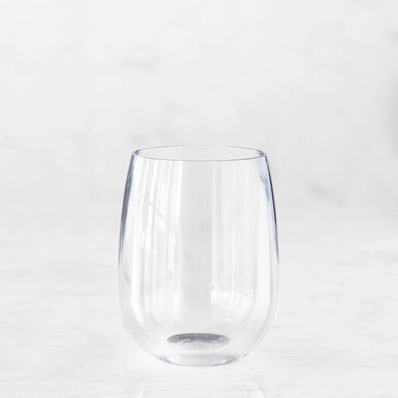 Clear glass product photograph of a stemless wine tumbler centered on a white marble surface featuring long vertical light reflections and a smooth rounded base against a bright minimalist background