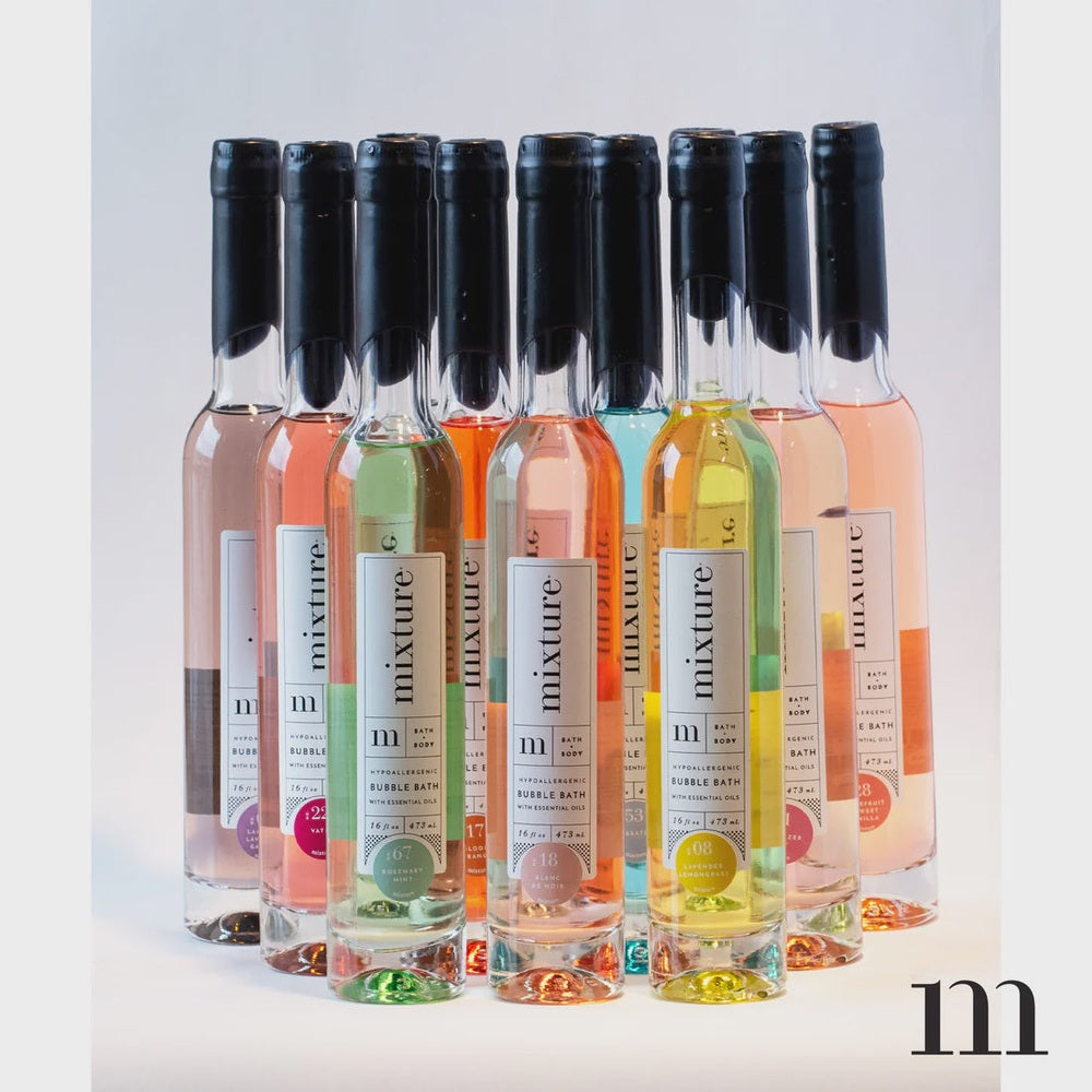 Commercial product photography for Mixture showcasing a collection of tall glass bubble bath bottles in a studio setting featuring vibrant liquids in lime green, sunset orange, and pale pink with minimalist white labels and sleek black neck seals