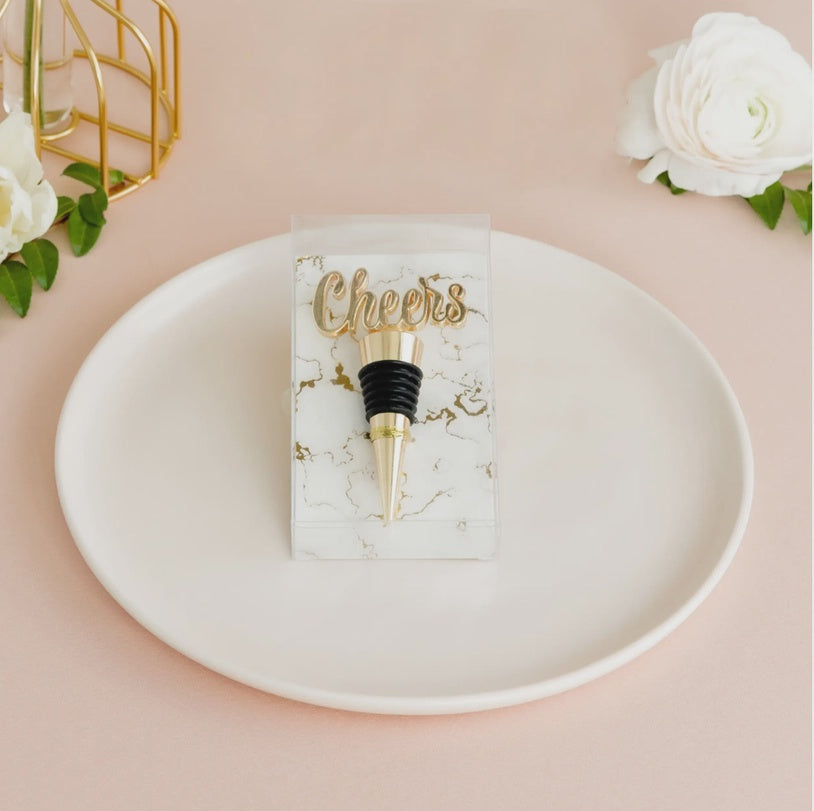 Lifestyle photograph of a metallic gold Cheers script wine bottle stopper on a white ceramic plate featuring a marble-patterned backing card and elegant white flowers on a soft pink surface