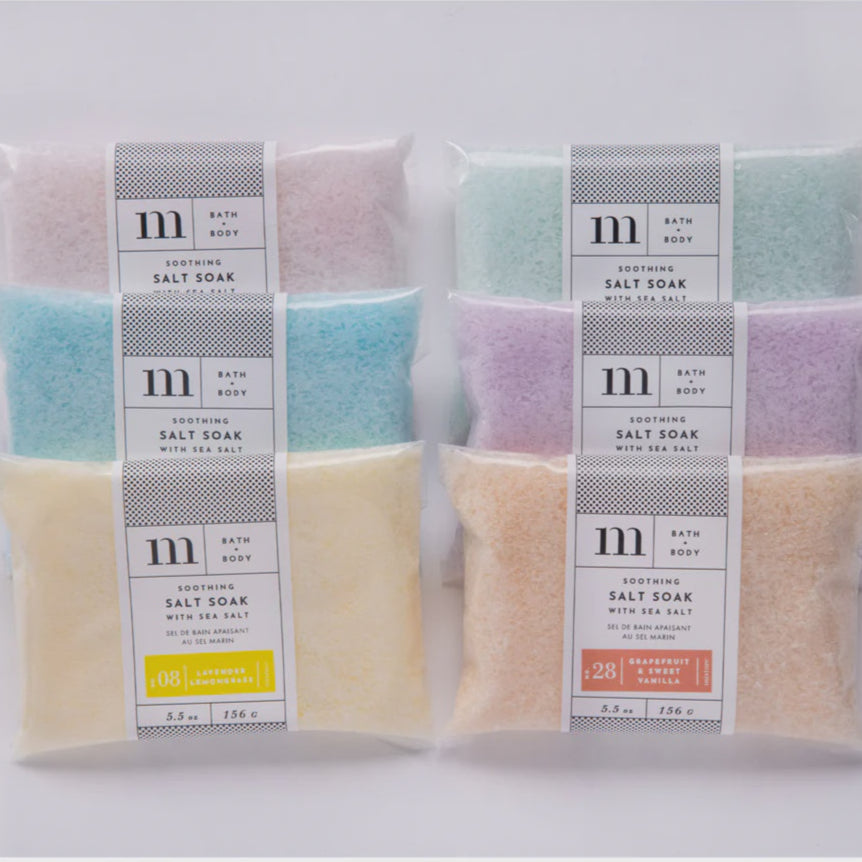 Commercial product photograph by m. bath body showcasing six transparent pouches of soothing salt soak on a white background, featuring the minimalist m logo and crystalline salts in pastel shades with labels for lavender lemongrass and grapefruit sweet vanilla