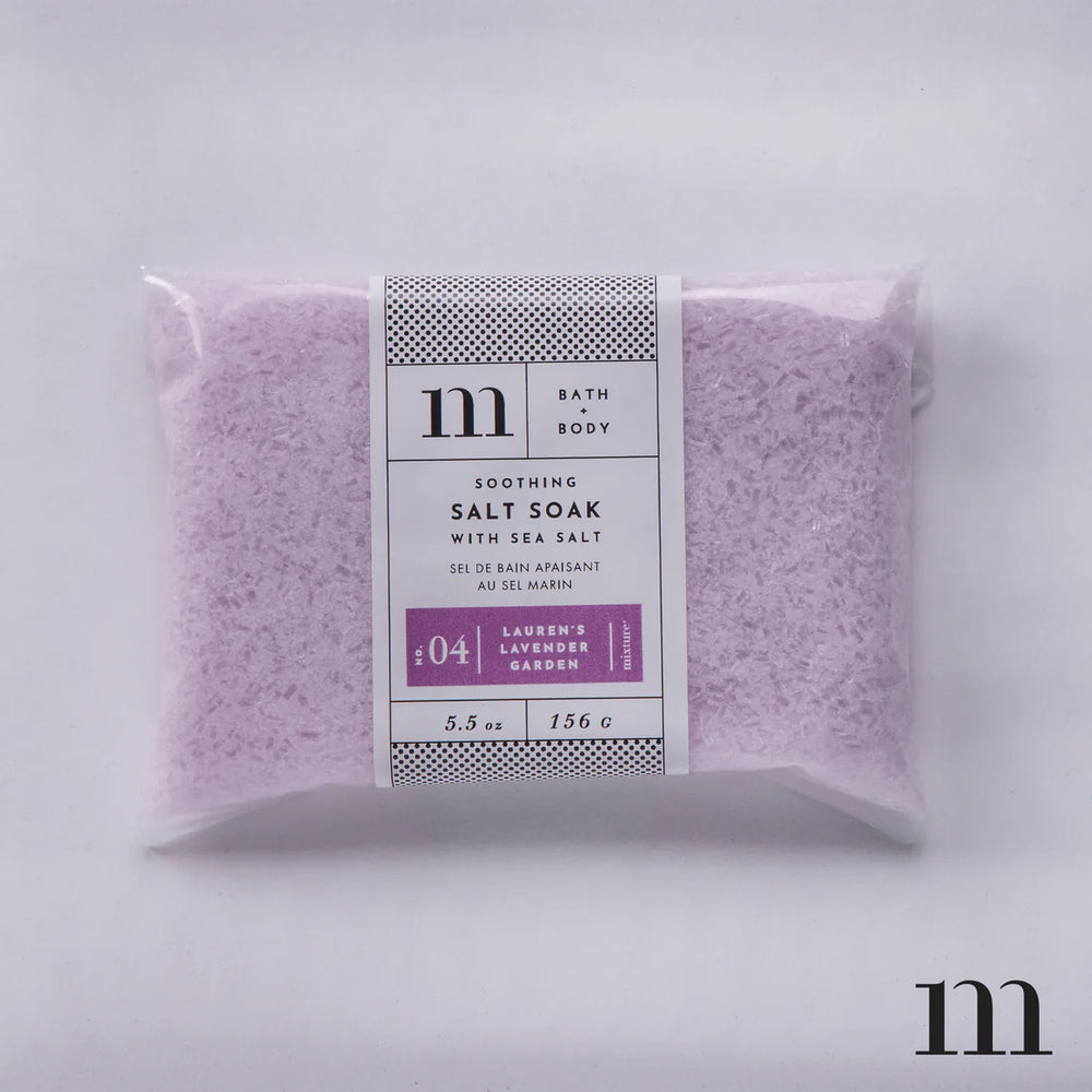 Product photography by Mixture of a pouch of Lauren's Lavender Garden Soothing Salt Soak on a neutral background featuring the brand's iconic lowercase m logo, pale purple salt crystals, and a white label with decorative dotted borders