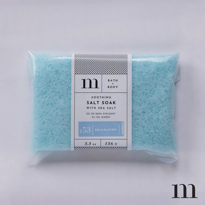 Studio product photograph by Mixture featuring a clear bag of No. 53 Relaxation Soothing Salt Soak filled with pale blue sea salt crystals and a white label with a black polka dot border and the m brand logo against a plain white background
