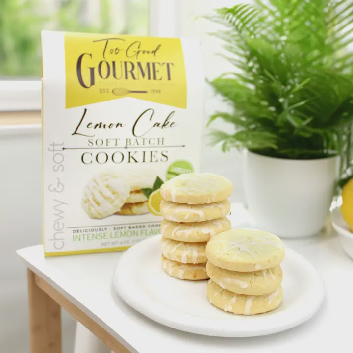Product photograph by Too Good Gourmet of lemon cake soft batch cookies stacked on a white plate in a bright indoor setting featuring white-iced glaze drizzles, a vibrant yellow branded box, and a lush green potted fern