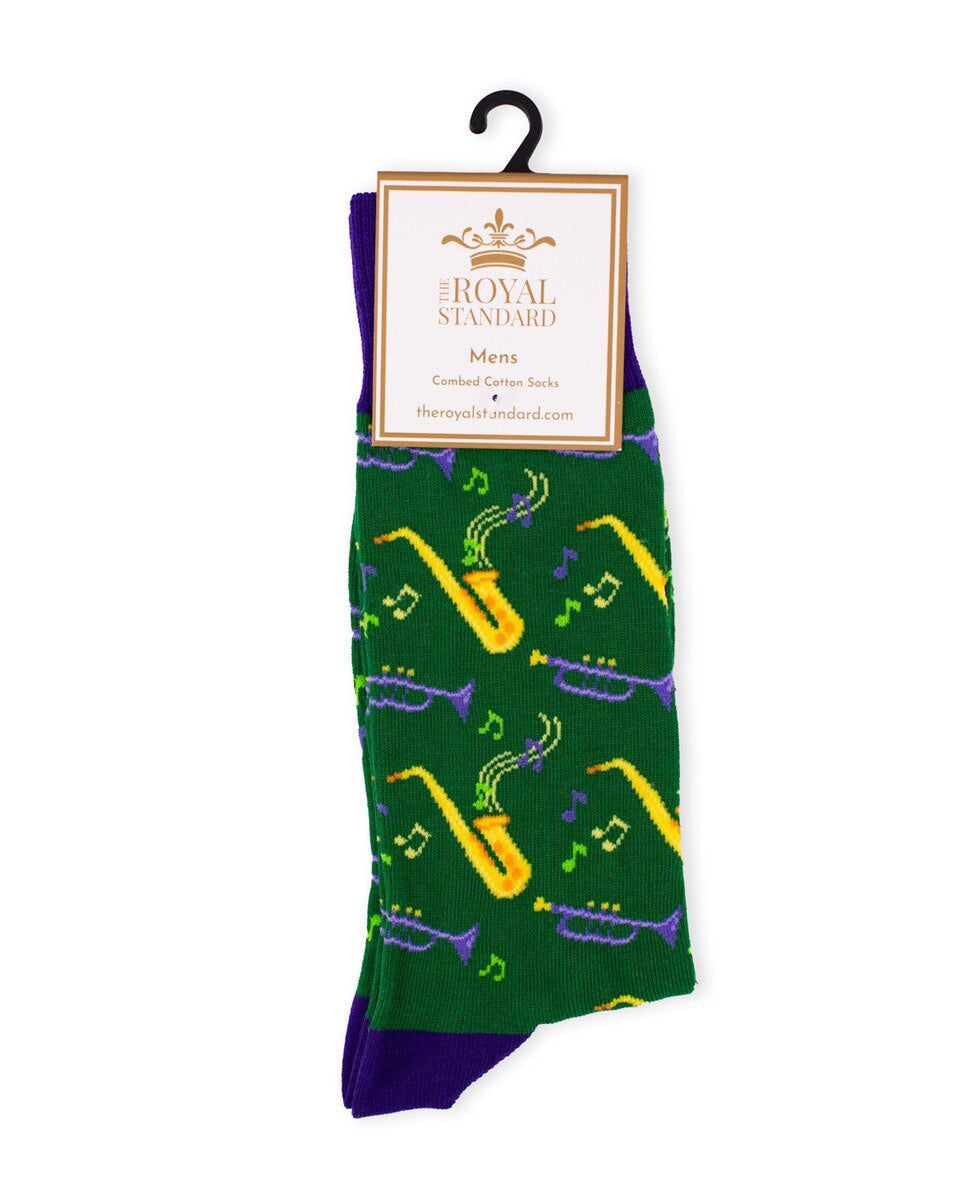 Product photography by The Royal Standard features green Mardi Gras themed men's socks with New Orleans jazz iconography including yellow saxophones, purple trumpets, and scattered musical notes