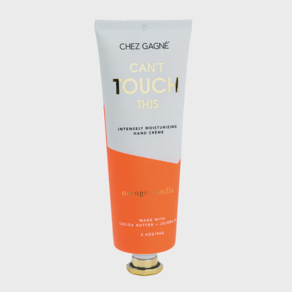 Product photograph by Chez Gagné of an orange vanilla hand cream tube featuring a vibrant orange and white color-blocked design, metallic gold lettering for the phrase Can't Touch This, and a gold hexagonal cap against a neutral background