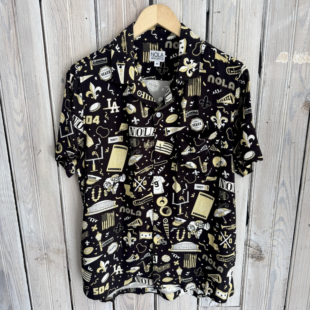 Photograph of a NOLA Mercantile short-sleeve button-down shirt featuring a black and gold New Orleans-themed pattern with the Caesars Superdome, Bourbon Street signs, fleur-de-lis symbols, and jazz instruments hanging against a rustic wooden wall