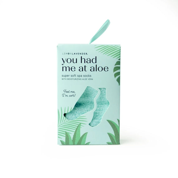 Commercial product photograph by Lemon Lavender featuring a mint green box of moisturizing spa socks on a white background with a foot-shaped cutout, tropical aloe plant illustrations, and a matching teal hanging ribbon