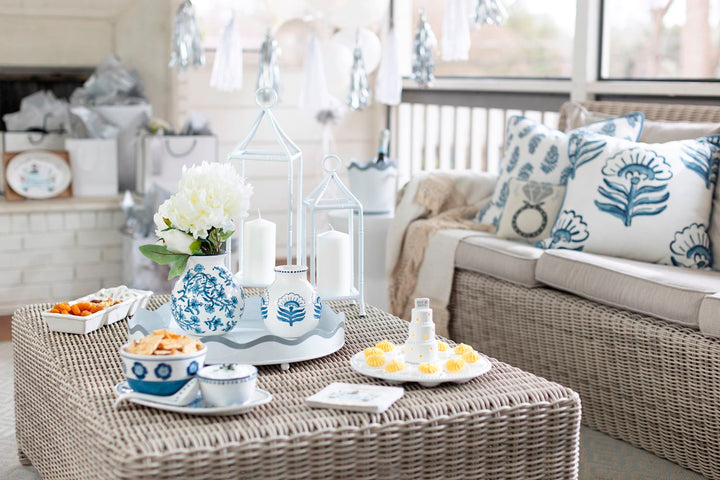 Photograph of a bridal shower celebration on a sunroom porch featuring blue and white chinoiserie vases with white peonies, a miniature wedding cake topper with yellow meringues on a wicker coffee table, and decorative pillows with a diamond ring motif