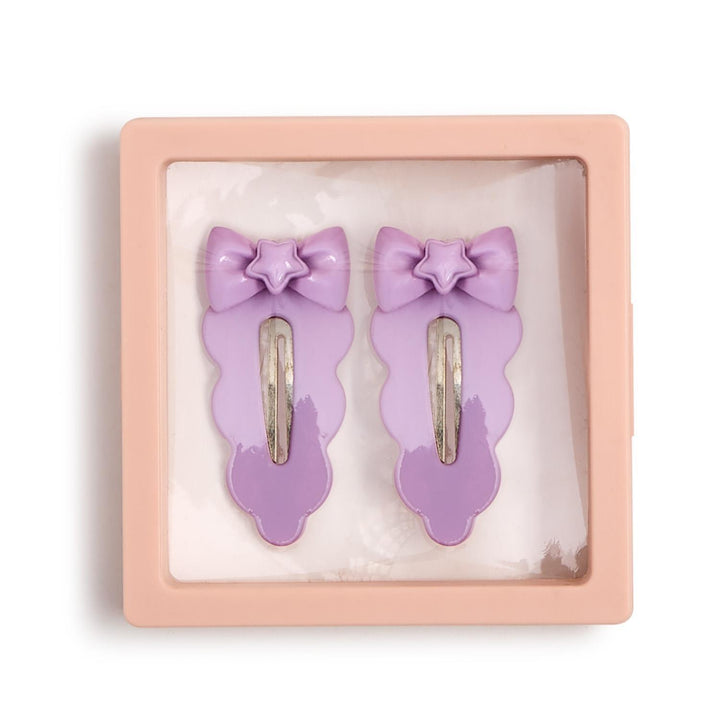 Product photograph of two lavender hair clips with star-centered bows and scalloped edges displayed inside a peach-colored square display box featuring silver snap-on fasteners