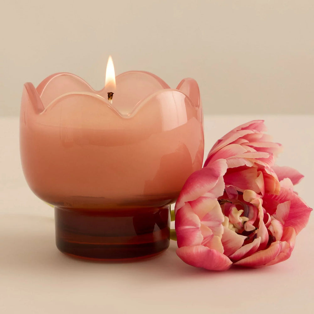 Still life photograph of a pink glass candle with a warm flickering flame and scalloped rim resting on a white surface next to a vibrant pink and cream ruffled tulip flower
