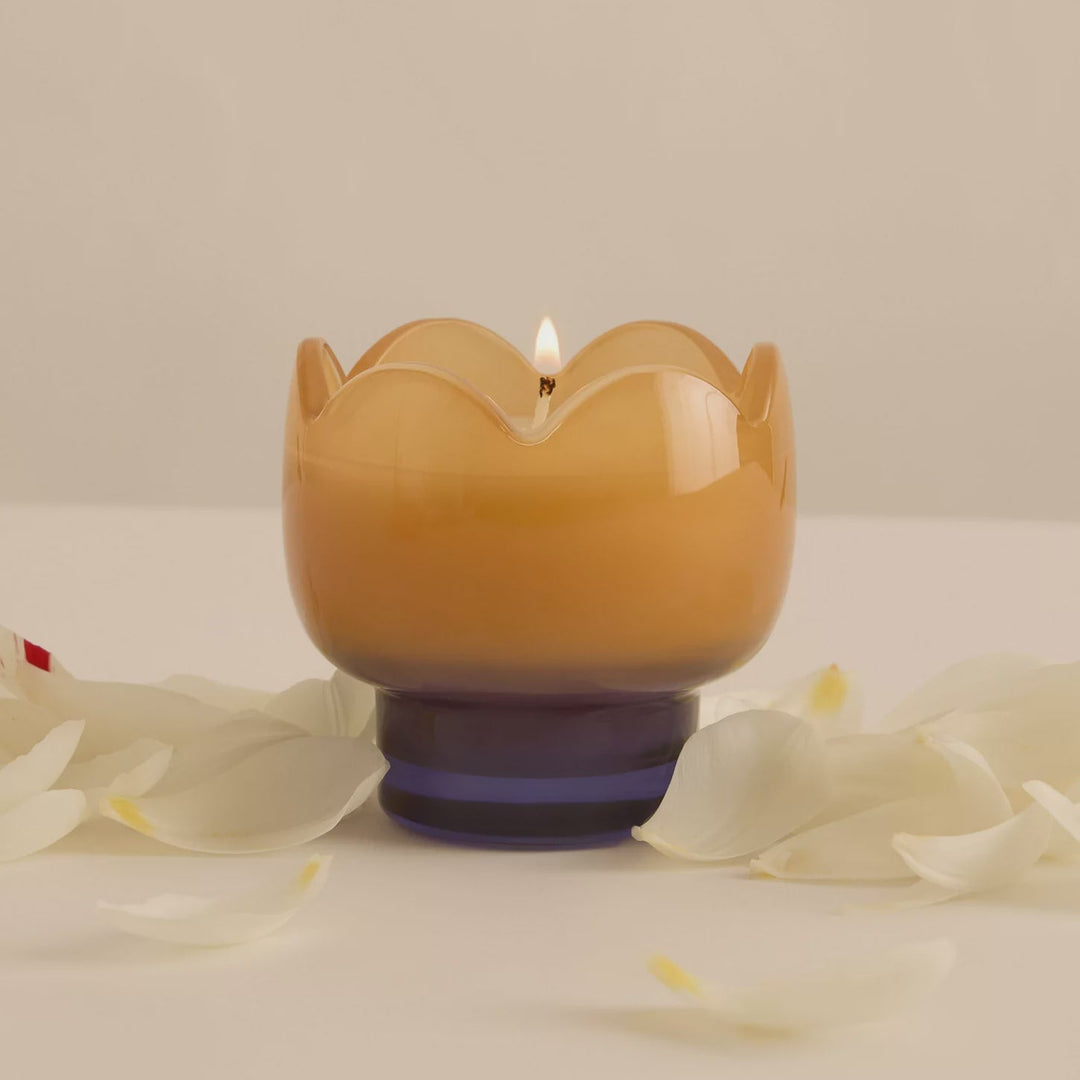 Photograph of a two-toned glass candle holder with a scalloped tulip-shaped rim, featuring a glowing amber top and deep indigo base surrounded by scattered white petals on a minimalist studio surface