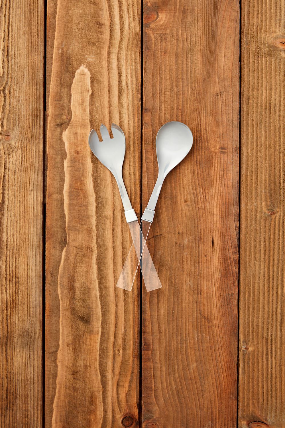 Digital photograph of a modern salad serving set on a rustic wooden surface featuring polished stainless steel heads and transparent handles arranged over textured vertical planks with rich grain and warm brown tones