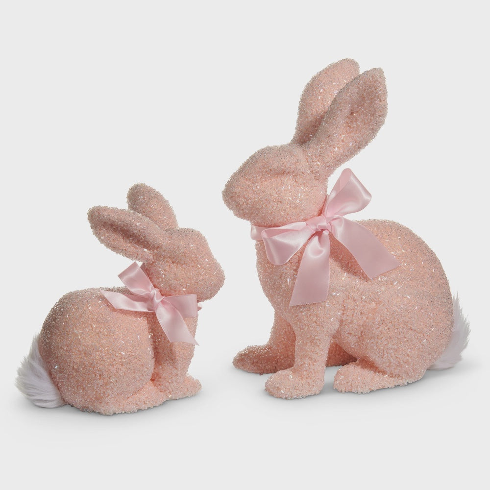 Decorative figurines of two seated pink bunnies on a plain white background featuring a shimmering tinsel-textured surface, matching satin neck ribbons, and soft white pom-pom tails