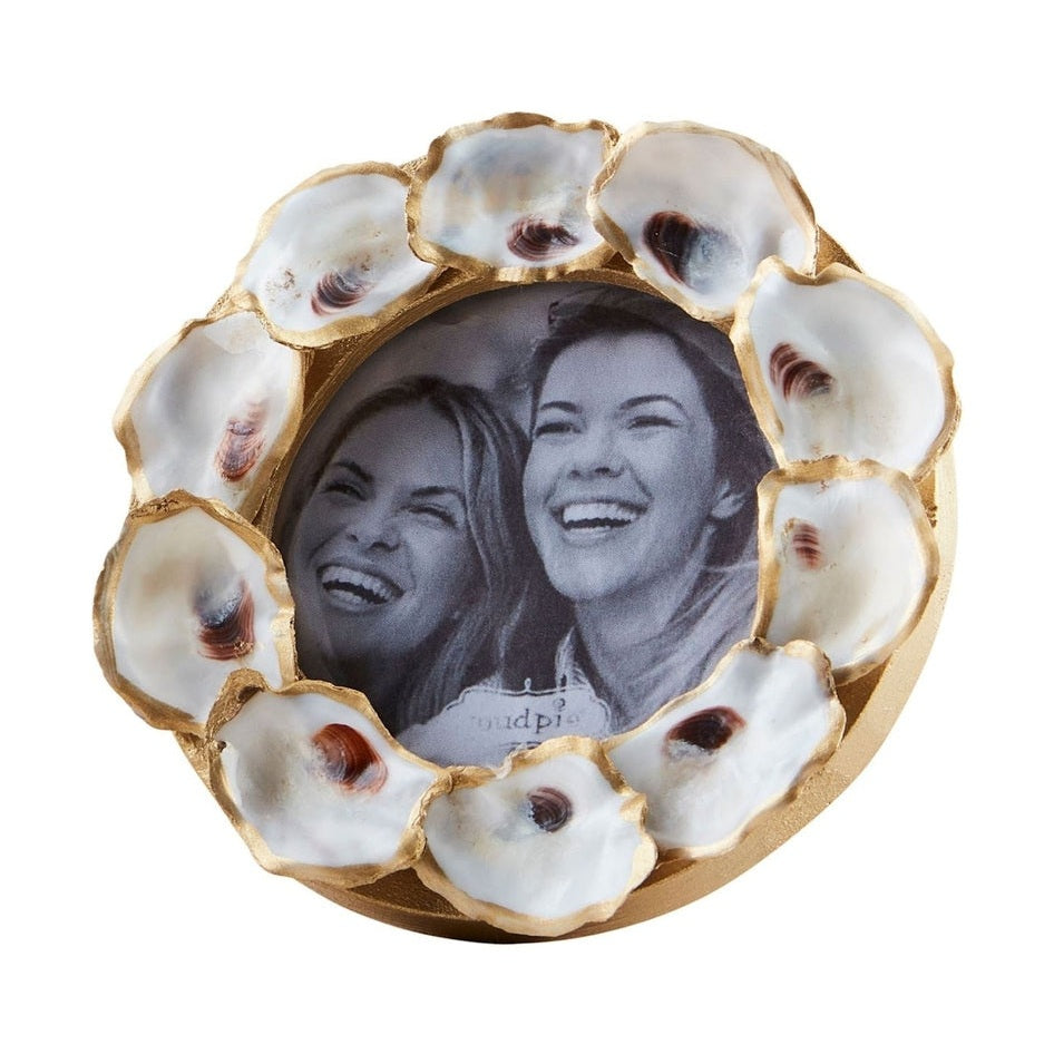 Mud Pie decorative oyster shell picture frame featuring a circular arrangement of shells with metallic gold-leaf edges and realistic brown muscle scars surrounding a black and white portrait of two laughing women