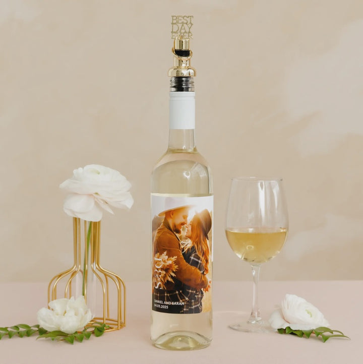 Product photography of a personalized wine bottle featuring a custom photo label and a gold Best Day Ever topper on a beige tabletop, styled with white ranunculus flowers in a gold wire vase and a filled wine glass