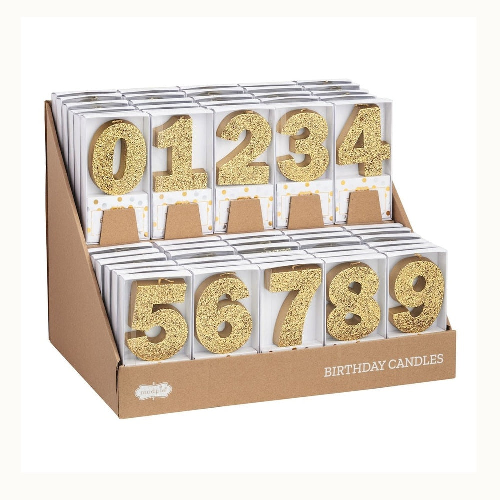 Product photograph of Mud Pie birthday candles featuring sparkling gold glitter numbers zero through nine arranged in a tiered kraft paper display stand with white boxes decorated in gold foil polka dots