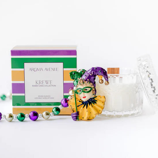 Product photograph for Aroma Avenue featuring the Krewe Mardi Gras Collection candle in a faceted glass jar, displayed alongside a purple, green, and gold striped box, a detailed ceramic harlequin mask, and a strand of metallic festive beads