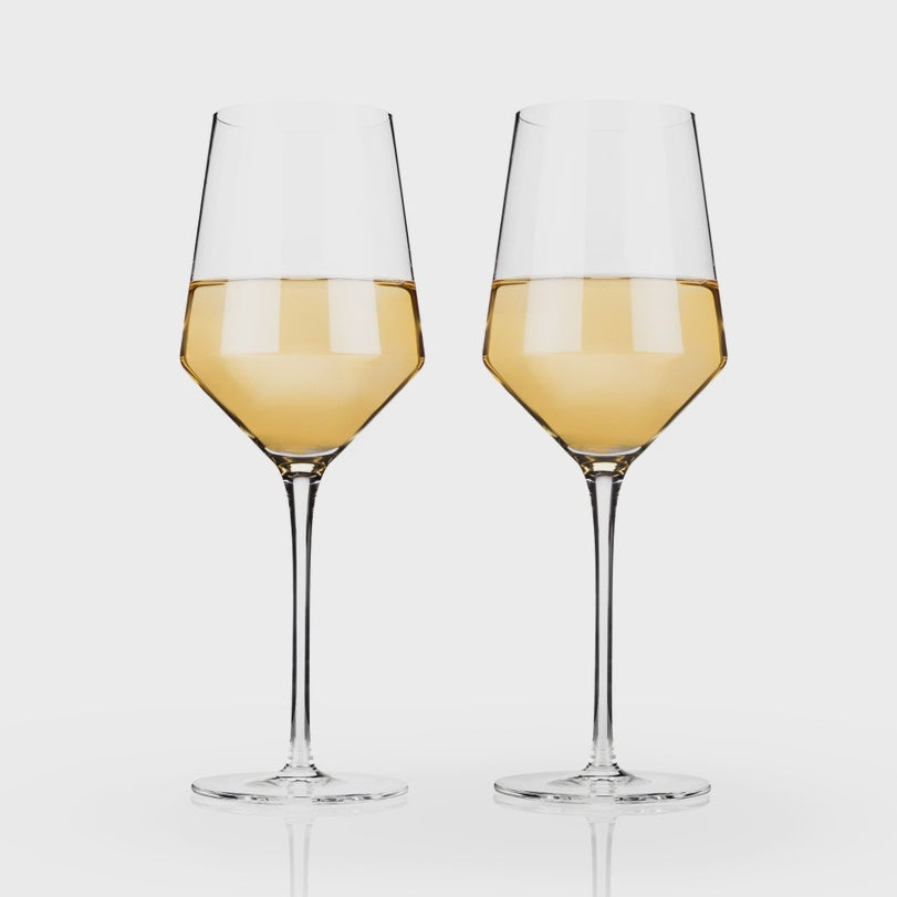 Studio photograph of two elegant crystal glasses filled with golden white wine featuring long slender stems and sharp angular bowls on a light grey background