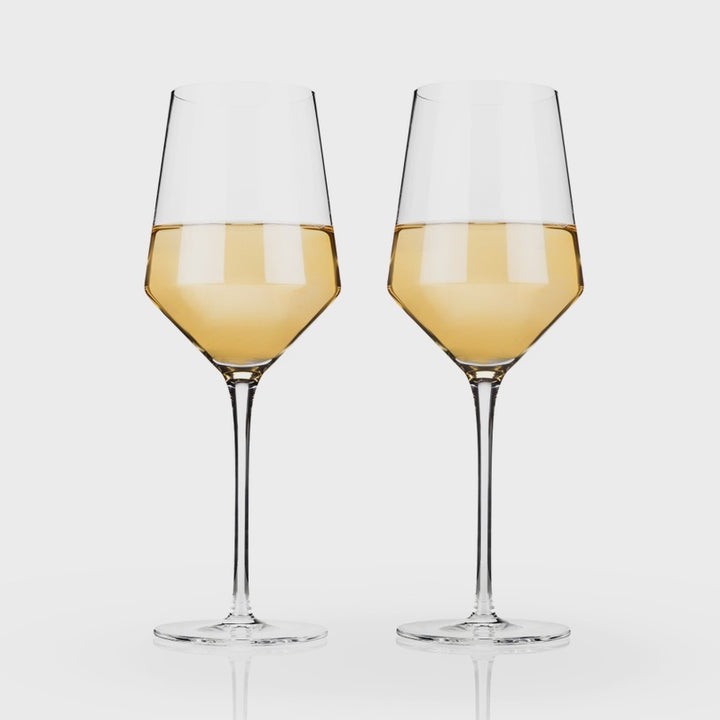 Studio photograph of two elegant crystal glasses filled with golden white wine featuring long slender stems and sharp angular bowls on a light grey background
