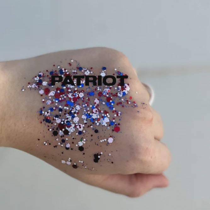 Cosmetic glitter body art on the back of a hand featuring red white and blue hexagonal flakes with shimmering particles and bold black text reading PATRIOT