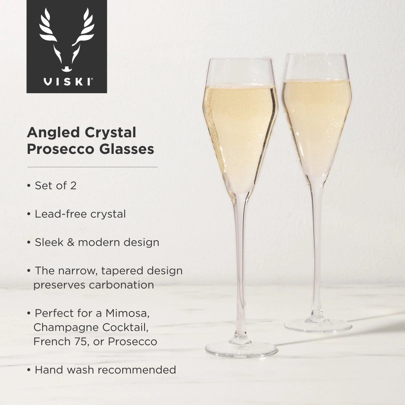 Commercial product photography by Viski featuring two angled crystal prosecco glasses filled with golden bubbling liquid on a white marble surface highlighting the Viski brand logo, slanted rims, and long slender stems
